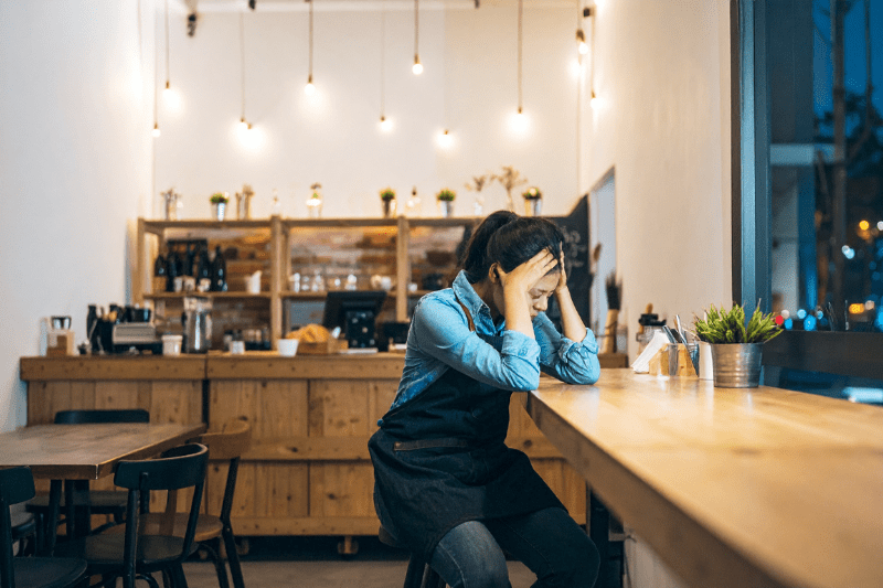A woman café owner sits alone at a table in a small, minimalist café at night, holding her head in frustration. The warm, dim lighting and clean interior emphasize a quiet, tense atmosphere, with empty tables around her and a dark street visible through the window.