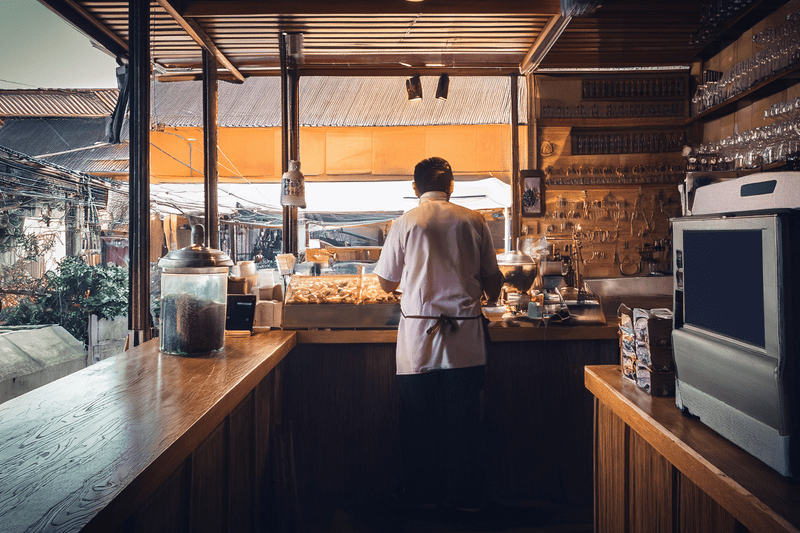 A man in a white apron stands behind the counter of a small café or bakery, facing a window that looks out onto a quiet urban alley. The interior is warm and rustic, with wooden countertops, glass jars, and stacked equipment, creating a cozy yet solitary atmosphere.