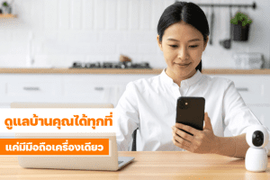 หญิงสาวกำลังดูหน้าจอมือถือในห้องครัว พร้อมกล้องวงจรปิดแบบสมาร์ตวางอยู่บนโต๊ะ แสดงถึงการควบคุมความปลอดภัยของบ้านผ่านแอปมือถือ